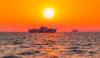 Wall Mural - Water trail foaming behind a passenger ferry boat in the Gulf of Izmir at red sun - Izmir, Turkey