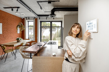 Young pretty woman controlling smart home devices with a digital tablet mounted on the wall. Wide angle view on stylish living room interior