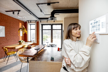 Young pretty woman controlling smart home devices with a digital tablet mounted on the wall. Wide angle view on stylish living room interior