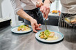 © AnnaStills - Close-up of assistant learning to decorate dish with salad with chef teaching him in kitchen