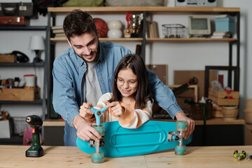 Cute little girl in eyeglasses using wrench while fixing wheels of blue skateboard while her father holding it and giving advice