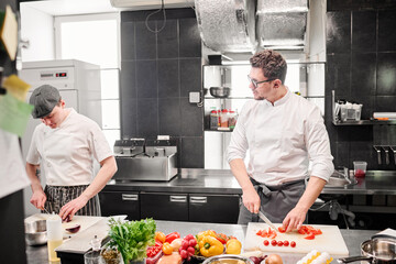 Young chef cutting tomatoes on cutting board and discussing menu with his assistant during their work at table in kitchen