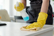 Young woman in protective yellow rubber gloves wiping dust on desk while spraying detergent and cleaning furniture in office