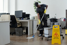 Contemporary Young Black Man In Workwear Cleaning Floor In Openspace Office In Front Of Yellow Plastic Signboard With Caution
