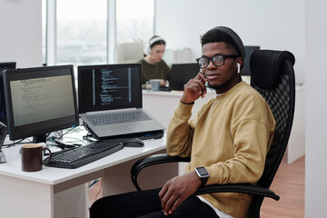 Poster - Young confident black man in casualwear with earphones sitting by workplace with compute monitor and laptop with decoded data