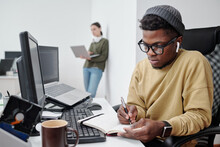 Serious African American Guy In Eyeglasses And Casualwear Making Notes In Copybook While Sitting By Desk In Front Of Computer