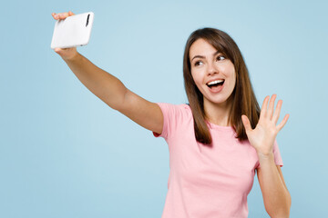 Wall Mural - Young smiling cheerful happy woman 20s wear pink t-shirt doing selfie shot on mobile cell phone post photo on social network waving hand isolated on pastel plain light blue background studio portrait