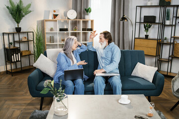 Wall Mural - Retired couple using laptop together sitting on sofa, smiling senior middle aged man and woman read internet news talking shopping online at home, elderly family customers with computer lifestyle