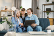 © sofiko14 - Family leisure, technology concept. Young grandson hugging his grandmother and grandfather in casual domestic clothes, sitting together on soft couch at home and watching interesting movie on laptop
