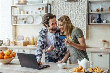 Happy young european husband with stubble and wife shopping online and show credit card in modern kitchen