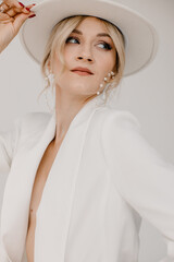 Wall Mural - Confident young blond woman smiling, looking at camera isolated on white background. Studio portrait of successful friendly female in white suit and hat, posing over white wall.