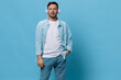 Cheerful happy tanned handsome man in casual basic t-shirt headphones look at camera hold hand on jeans pocket posing isolated on blue studio background. Copy space Banner Mockup. Music concept