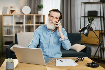 Wall Mural - Pleasant mature caucasian businessman talking on mobile and working on laptop at table. Senior male in casual wear sitting at workplace and using modern gadgets.