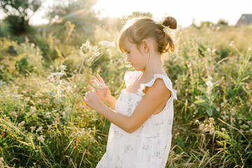 Adorable little girl wearing natural white dress with wildflower motiv in green field with wild carrot flowers at summer, outdoor lifestyle. Freedom concept