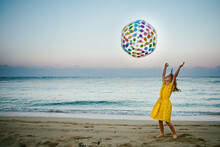 Blond Girl Throws Rainbow Ball Joyfully On Beach During Sunset
