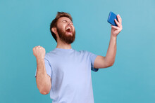 Portrait Of Extremely Happy Handsome Bearded Man Gamer Playing Video Game On Mobile Phone, Celebrating Victory, Complete Level. Indoor Studio Shot Isolated On Blue Background.
