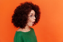 Profile Portrait Of Shocked Woman With Afro Hairstyle In Green Casual Style Sweater Standing With Open Mouth, Looking Away, Sees Something Surprising. Indoor Studio Shot Isolated On Orange Background.