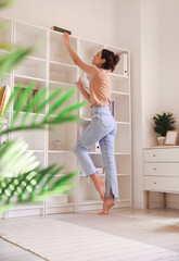 Poster - Young barefoot woman taking book from shelf at home