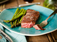 Strip Steak With A Piece Pierced On A Fork; Served With Asparagus On A Blue Plate