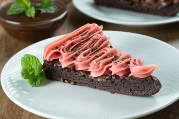 Wall Mural - Piece of Chocolate Brownie Slice with Strawberry and Chocolate Icing. Wooden table with mint and chocolate chips in the background