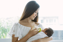 Portrait Of Beautiful Young Asian Mother Hold Bottle Milk For Feeding Her Newborn Baby And She Feels Happiness At Home. Happy Motherhood Concept.