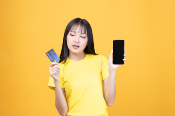 Wall Mural - Portrait Asian happy young girl smiling cheerful and showing plastic credit card while holding mobile phone isolated on yellow background