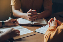 Christians And Bible Study Concept.Christian Family Sitting Around A Wooden Table With Open Bible Page And Holding Each Other's Hand Praying Together.