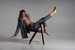A model girl poses professionally for a photographer on a chair in complex acrobatic recumbent poses. In a formal suit and a sexy unbuttoned gray jacket, barefoot