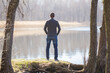© fotoduets - Young adult man in dark clothes standing at lake shore between trees and looking far away. Thinking about life. Peaceful atmosphere in sunny spring morning. Spending time alone in nature. Back view.