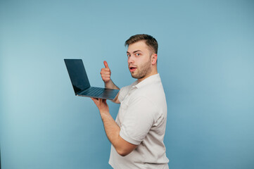 Adult man with bristles in a white T-shirt with a laptop in his hand on a blue background shows a gesture like and looks at the camera.