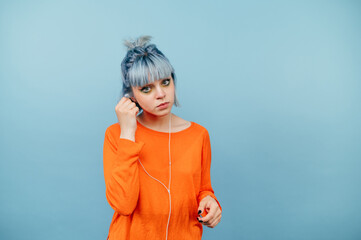 Wall Mural - Beautiful hipster girl in a green sweater and colored hair and listens to music in headphones with a serious face on a blue background