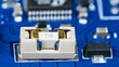 Small electrical fuse in white holder and black linear fixed voltage regulator on blue PCB. Printed circuit board detail with surface mount technology of electronic components and bokeh in background.