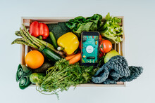 Top View Wooden Box With Fresh Vegetables And Phone With Active Online Mobile Application With Healthy Diet Program On The White Background. Healthy Dieting, Weight Loss, Healthy Food Delivery Concept