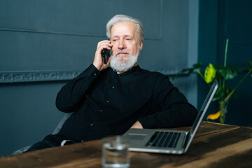 Wall Mural - Thinking gray-haired mature aged business man talking on mobile phone sitting at wooden table with laptop computer. Portrait of serious senior older male having conversation on smartphone.