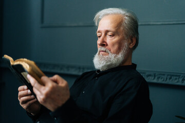 Wall Mural - Close-up side view of senior gray-haired aged male reading paper book sitting on chair in dark living room, relax and rest with literature. Handsome mature adult man reading book at home alone.