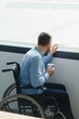 © Serhii - Disabled Person Sits in Wheelchair Against Window. Serious Sad Caucasian Man Wearing Casual Clothes and Look at Large Panoramic View in Bright Modern Living Room or Hospital.