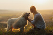 Happy young woman is sitting on the hill at sunset lovingly hugging her large breed dog.