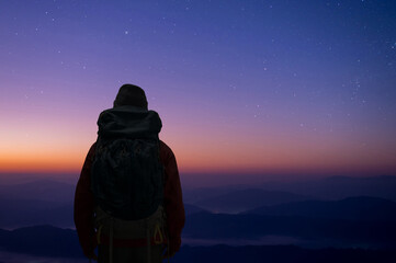 Wall Mural - Silhouette of young traveler  with backpack standing alone on top of the mountain watched beautiful night sky, star and milky way over the sky with sunlight at twilight time.