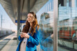 © Big Shot Theory - Young woman waiting at rail station. Holding smartphone and waiting for her train. Public transport.