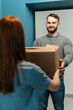 © dikushin - Vertical shot of friendly bearded courier male delivering cardboard box parcel to female customer on doorway at home. Back view of young woman client receiving parcel package post from delivery man.