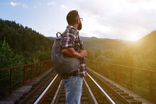 A Young Tourist With A Beard And Glasses In A Plaid Shirt With A Backpack Stands On The Railway Tracks And Looks Into The Distance At The Mountains.