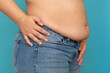 © Юля Бурмистрова - Side view of unrecognizable fat plump obese overweight woman stand in blue jeans putting hand on waist, showing excess naked belly on blue background. Body positive, obesity, weight loss, liposuction.