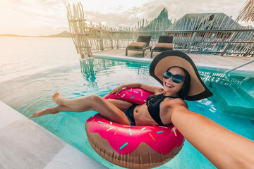 Relaxing woman floating in donut inflatable swimming pool toy at luxury resort taking selfie photo using sunbathing. Travel vacation hotel lifestyle