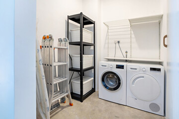 Interior of a storeroom with modern washing machine, dryer machine, foldable ladder, shelf with tools and boxes.