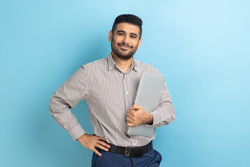 Portrait of confident businessman standing holding closed laptop or folder, has positive expression, keeps hand on hip, wearing striped shirt. Indoor studio shot isolated on blue background.