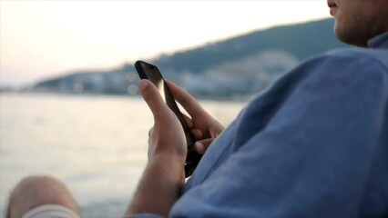 Close up of a young man is using his smartphone while sitting at the beach near the sea.