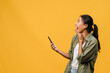 © tirachard - Surprised young Asia lady using mobile phone with positive expression, smiles broadly, dressed in casual clothing and stand isolated on yellow background. Happy adorable glad woman rejoices success.