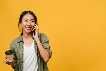 Wall Mural - Young Asia lady talk by phone and hold coffee cup with positive expression, smile broadly, dressed in casual cloth feeling happiness and stand isolated on yellow background. Facial expression concept.