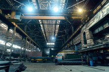 Large Girder Or Beam Crane Transports Iron Pipes Inside Large Dark Workshop In Metallurgical Factory.