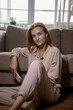 © Мария Чичина - Caucasian young good looking woman with red hair sitting on the floor near the sofa in room and tapping or playing on the smartphone.